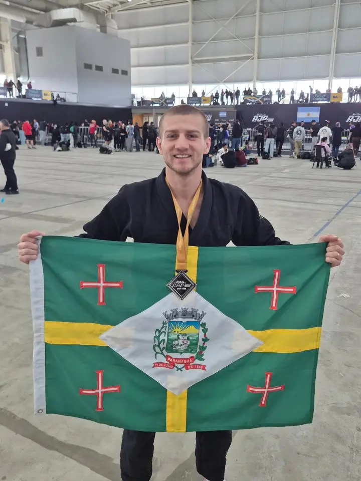 Gabriel sorri de quimono preto, com a medalha no pescoço, segurando aberta a bandeira de Paranaguá — verde, com cruz amarela e o brasão num losango branco — num grande pavilhão com público e tatames, no AJP Tour Argentina 2026.