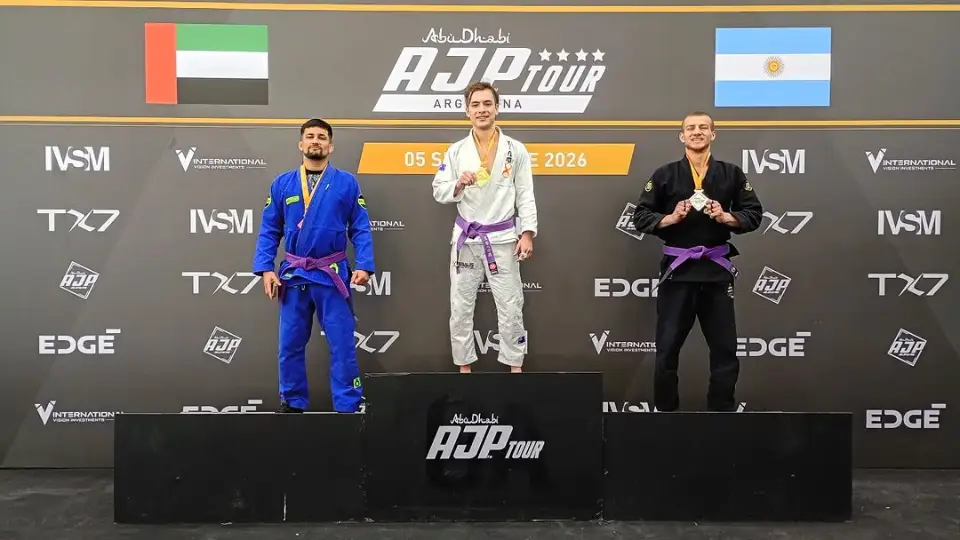 Pódio do AJP Tour Argentina 2026, com as bandeiras dos Emirados Árabes e da Argentina no alto do painel. Três atletas de faixa roxa: no centro, de quimono branco, o 1º lugar; à esquerda, de quimono azul; à direita, no degrau do 3º lugar, Gabriel, de quimono preto, com a medalha.