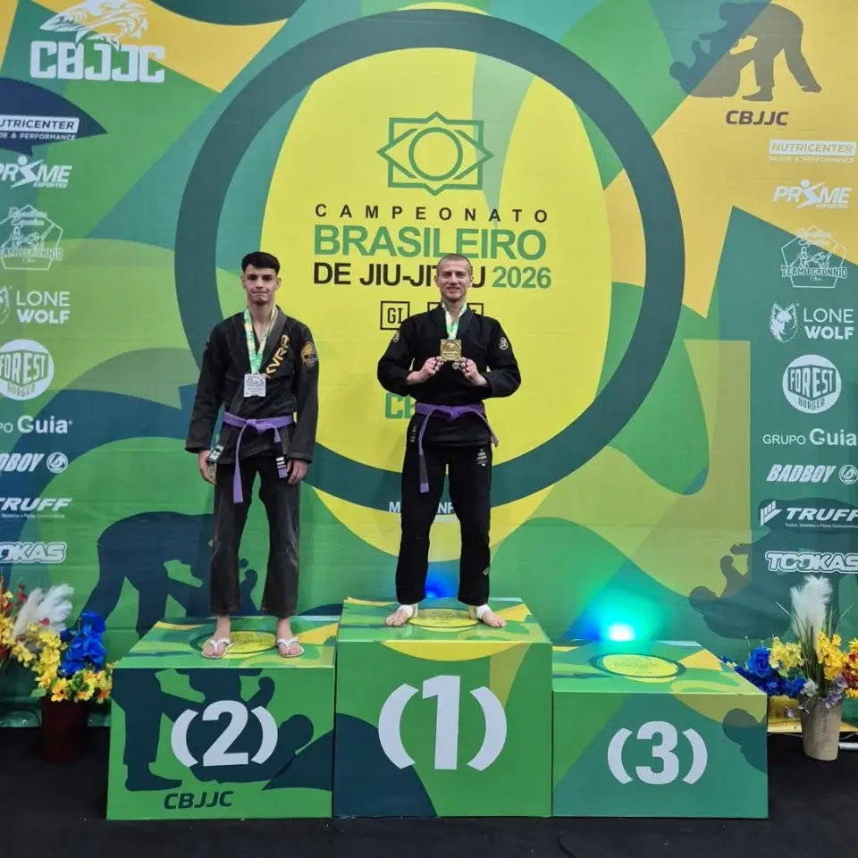 Pódio verde e amarelo do Campeonato Brasileiro de Jiu-Jitsu 2026 (CBJJC). Gabriel, de quimono preto e faixa roxa, está no degrau mais alto, o número 1, mostrando a medalha de ouro. Ao lado, no degrau 2, outro atleta de quimono preto.