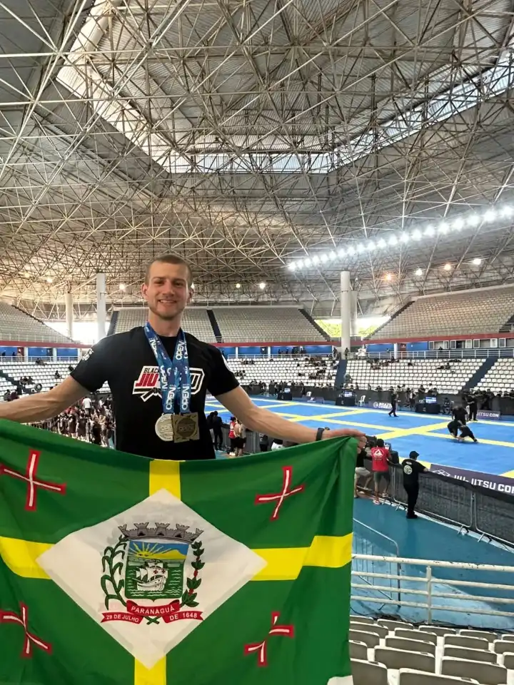 Gabriel, de camiseta preta e com duas medalhas no pescoço, estende a bandeira de Paranaguá — verde, com uma cruz amarela e o brasão da cidade num losango branco — dentro do ginásio do campeonato em Manaus, com tatames azuis ao fundo.