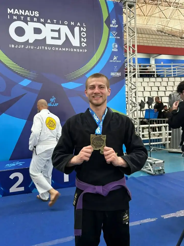 Gabriel sorri de quimono preto e faixa roxa, segurando uma medalha com as duas mãos, na frente do painel do Manaus International Open 2026. Ao fundo, o pódio e outros atletas.