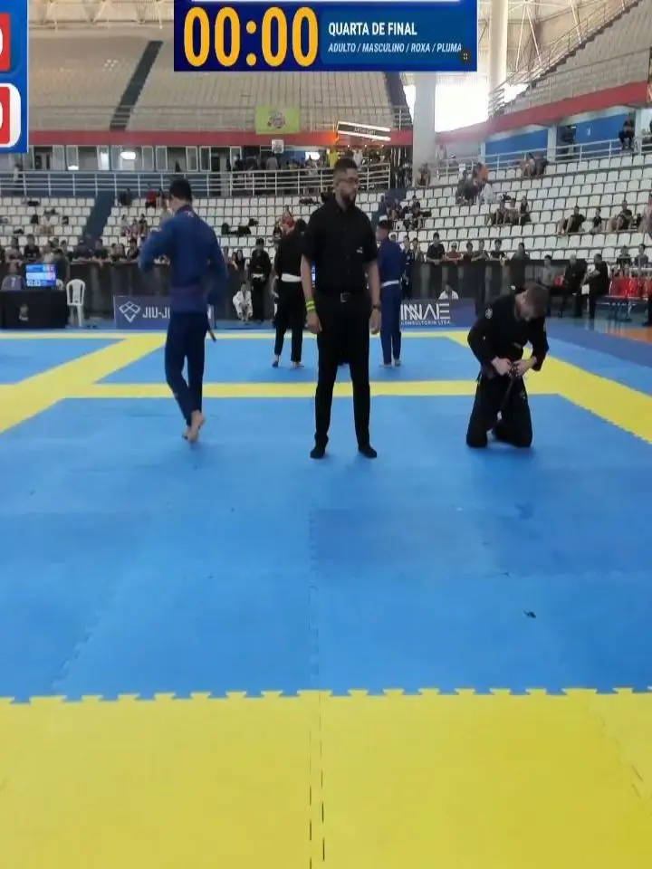 Tatame azul e amarelo do Manaus International Open 2026 durante uma quarta de final. No centro, o árbitro de preto; um atleta de quimono preto está ajoelhado ajeitando a faixa e outro, de quimono azul, caminha. No placar, "00:00 — Quarta de final".