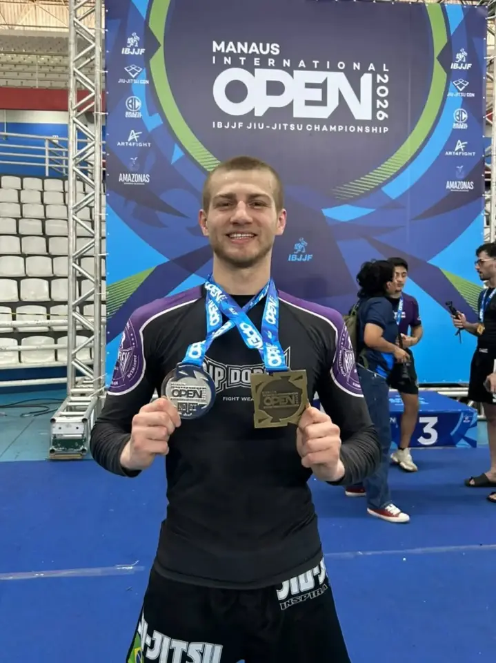 Gabriel sorri de frente, de camiseta de lycra preta e roxa, segurando duas medalhas penduradas no pescoço. Atrás dele, o painel azul do Manaus International Open 2026 (IBJJF) e o ginásio com arquibancadas.